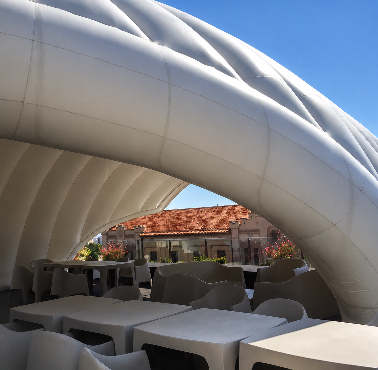 Close view beneath the white inflatable roof, where rounded membrane ribs span above terrace tables and chairs in bright daylight.