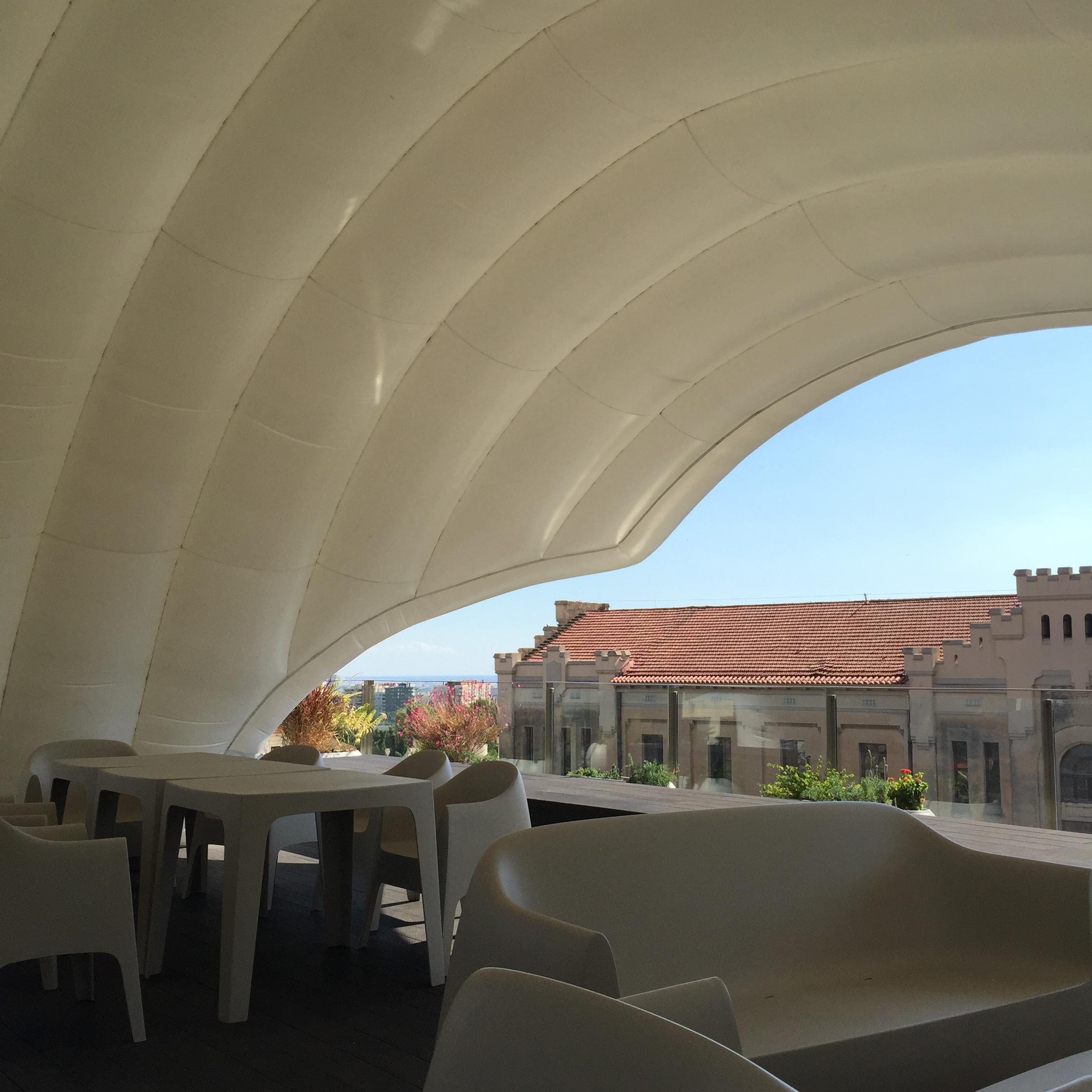 A white inflated membrane canopy arches above terrace tables and chairs, framing blue sky and a tiled Barcelona roofline.