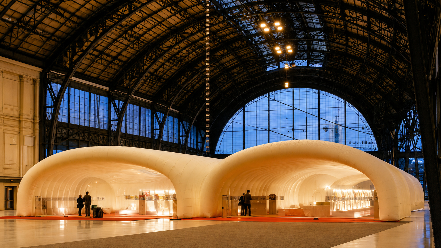 Two connected white inflatable pavilions glow inside a dark iron-and-glass hall, with visitors standing at their open entrances.